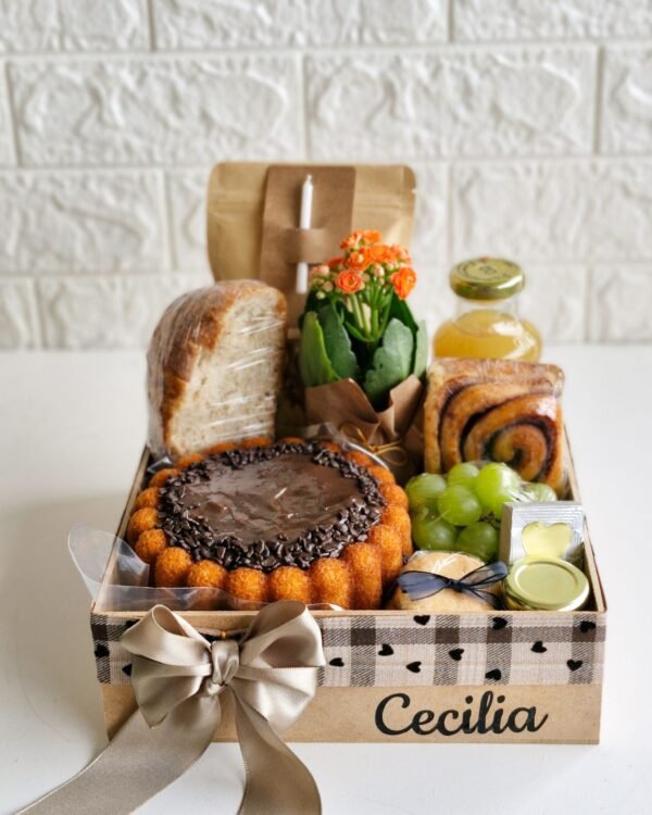 Cesta Feliz Dia 3 Cesta de aniversário em caixa de MDF decorada com fita xadrez bege com pequenos corações pretos e laço de cetim dourado suave. O nome 'Cecilia' está aplicado em preto. A cesta farta inclui bolo de cenoura com brigadeiro, flores Kalanchoe laranjas, pão artesanal, suco, uvas, rolinho de canela, Polenguinho, geleia, bem-vivido com laço preto e vela de aniversário.