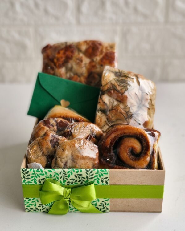 Cesta Encanto 4 Cesta de café da manhã farta em caixa de MDF decorada com estampa de folhas verdes e laço de fita de gorgurão verde claro. Acompanha envelope verde escuro, focaccias rústicas de fermentação natural, cookies abundantes com gotas de chocolate e rolinhos de canela.