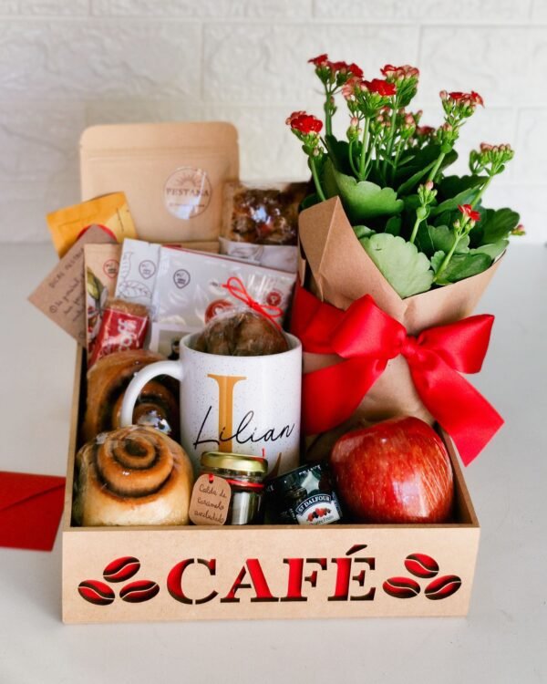 Cesta Café 1 Cesta de café da manhã premium em caixa de MDF com a palavra 'CAFÉ' em fundo vermelho. Destaque para uma caneca branca personalizada com o nome 'Lilian', uma maçã vermelha, flor Kalanchoe vermelha com laço de cetim elegante, geleia, potinho com tag 'calda de caramelo aveludado', pães artesanais e embalagem kraft com a logo da Pestana.