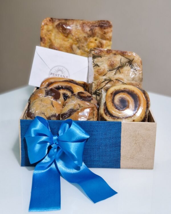 Inicio 8 Cesta de café da manhã em caixa de MDF envolta em tecido azul texturizado com um grande laço de fita de cetim azul bic. Exibe focaccias artesanais de provolone e alecrim, rolinhos de canela, cookies de chocolate e um envelope branco clássico com o selo da Pestana.