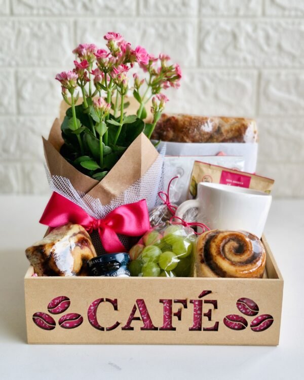 Cesta Café 3 Cesta de café da manhã em caixa de MDF vazada com a palavra 'CAFÉ' revelando estampa floral rosa. A composição inclui uma flor Kalanchoe rosa com laço de cetim pink, caneca branca, uvas verdes em embalagem transparente, rolinhos de canela brilhantes, geleia francesa St. Dalfour, pão artesanal e pacotes de café.