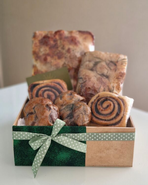 Inicio 7 Cesta de café da manhã em caixa de MDF decorada com faixa de tecido verde escuro texturizado e um laço de fita verde clara com bolinhas brancas (poá). Exibe fartura de pães rústicos, rolinhos de canela, cookies com grandes gotas de chocolate e um cartão pardo.