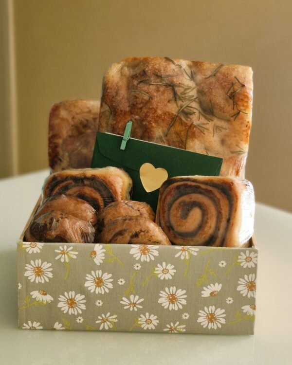 Cesta Encanto 5 Cesta de café da manhã em caixa de MDF forrada com tecido estampado de margaridas brancas em fundo verde sálvia. A composição farta inclui focaccias rústicas, cookies de chocolate, rolinhos de canela e um envelope verde escuro selado com adesivo de coração dourado e preso com mini pregador.