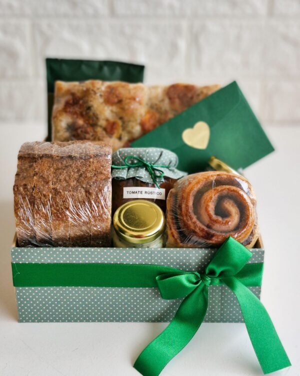 Cesta Partilhar 1 Cesta de café da manhã farta para duas pessoas em caixa de MDF forrada com tecido verde poá e laço de cetim verde escuro. O banquete inclui fatias grossas de pão 100% integral, focaccia de fermentação natural, rolinho de canela, um potinho com polpa de tomate rústico, patê e um envelope verde escuro com selo de coração dourado.