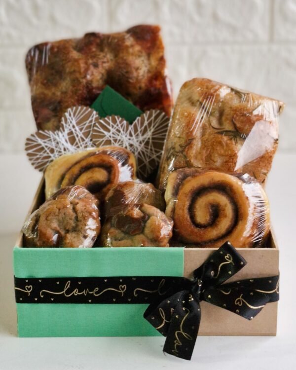 Cesta Encanto 1 Cesta de café da manhã em caixa de MDF com base verde água e laço preto escrito 'love' em dourado. Contém focaccia de provolone e alecrim, rolinhos de canela, mini cookies de chocolate, envelope verde escuro e tags decorativas em formato de coração vazado.