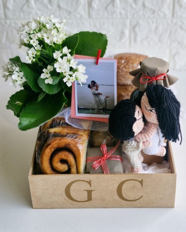 Cesta Taboa 1 Cesta de casamento em caixa de MDF crua com as iniciais 'G C' aplicadas em dourado. No interior, um casal de amigurumis em crochê abraçados em clima de romance, foto polaroid do casal presa a flores brancas, rolinho de canela, cookies e potes com acabamento em tecido rústico. Presente de luxo para noivos em São Gonçalo.