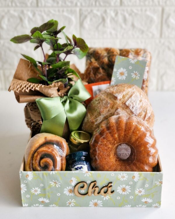 Cesta Chá da Tarde 3 Cesta de chá da tarde em caixa com estampa de margaridas verdes e letreiro de madeira escrito 'Chá'. A composição inclui um bolo caseiro com selo Pestana, pão rústico de fermentação natural, rolinho de canela, geleia francesa St. Dalfour, focaccia de alecrim e uma planta aromática em vaso decorado com laço de cetim verde. Presente afetivo em São Gonçalo.