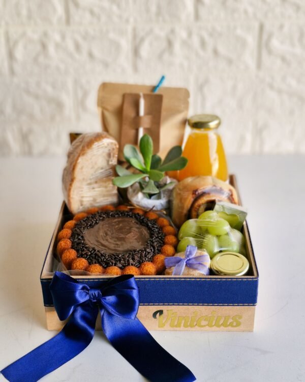 Cesta Feliz Dia 1 Cesta de café da manhã de aniversário em caixa de MDF com fita e laço azul marinho, decorada com o nome 'Vinicius' em dourado. A cesta é muito farta e contém um mini bolo de cenoura com cobertura de chocolate granulado, pão de fermentação natural, suco de laranja, rolinho de canela, uvas verdes, bem-vivido com laço lilás, potinho de vidro, planta suculenta e uma velinha branca fixada na embalagem do café.
