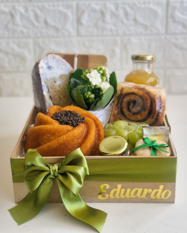 Cesta Partilhar 3 Cesta de café da manhã farta em caixa de MDF com fita e laço de cetim verde musgo. O nome 'Eduardo' aparece em dourado. No interior: bolo de cenoura em formato de flor com miolo de chocolate granulado, pão rústico de fermentação natural, flores brancas, suco de laranja, rolinho de canela, uvas verdes, Polenguinho, bem-vivido com laço verde e vela.
