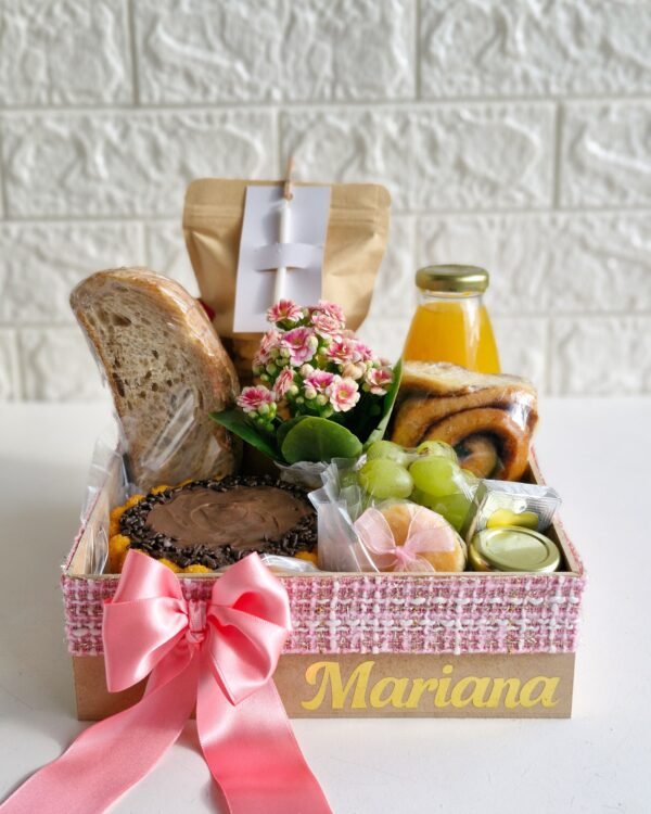 Cesta Feliz Dia 2 Cesta de café da manhã de aniversário em caixa de MDF decorada com fita texturizada rosa e laço de cetim rosa claro, exibindo o nome 'Mariana' em dourado. Contém mini bolo de cenoura com chocolate, flores Kalanchoe cor-de-rosa, pão rústico, rolinho de canela, suco de laranja, uvas, Polenguinho, potinho de vidro, bem-vivido com fita rosa e um drip coffee com vela de aniversário.