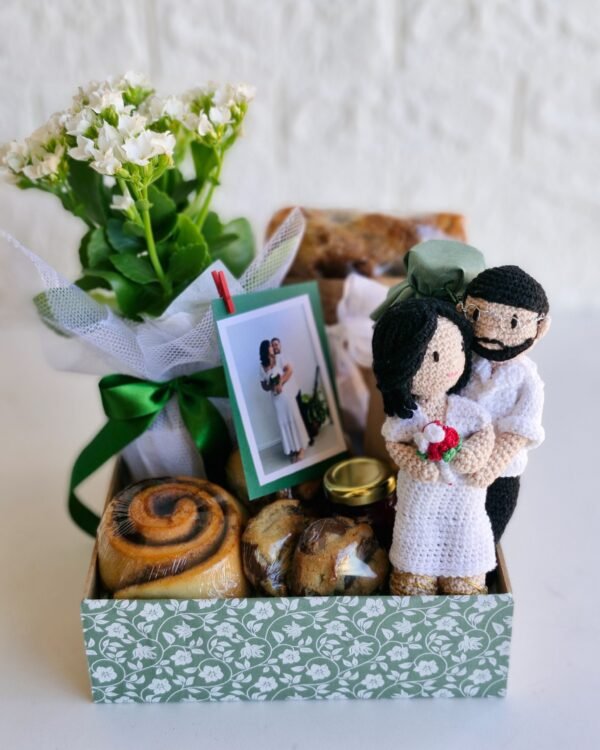 Cesta Infantil com Amigurumi 2 Cesta de casamento premium em caixa verde com estampa floral. Contém um casal de amigurumis vestidos de noivos (noiva de branco com buquê e noivo de óculos), flor natural Kalanchoe branca com laço verde, e uma foto polaroid do casal presa com mini pregador. Inclui também rolinho de canela e cookies. Presente para recém-casados em São Gonçalo.