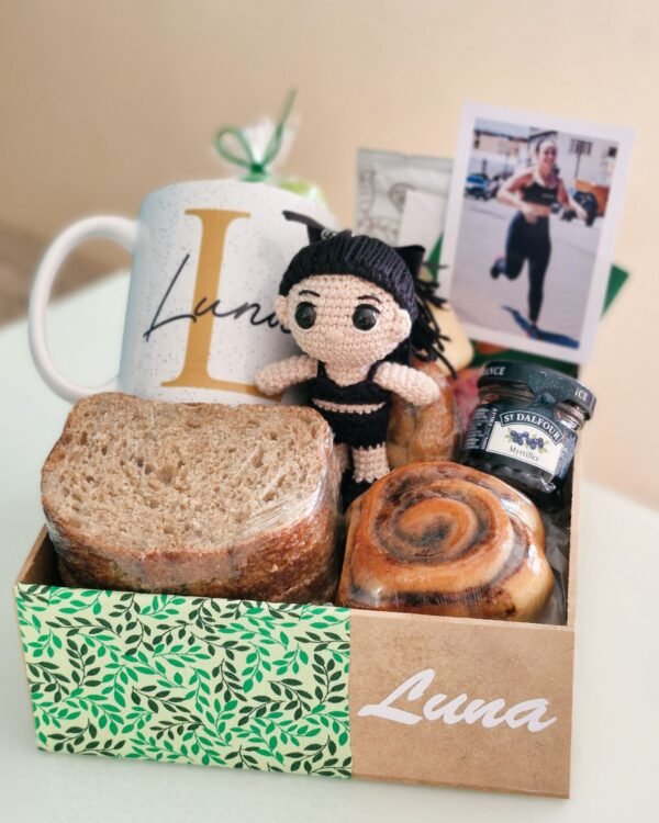Inicio 6 Cesta de café da manhã fitness premium em caixa de MDF com estampa de folhas verdes e o nome 'Luna'. A composição exclusiva exibe uma boneca amigurumi em crochê com roupas de ginástica pretas, foto polaroid de uma mulher correndo, caneca personalizada, fatias de pão 100% integral, rolinho de canela e geleia de mirtilo sem açúcar.