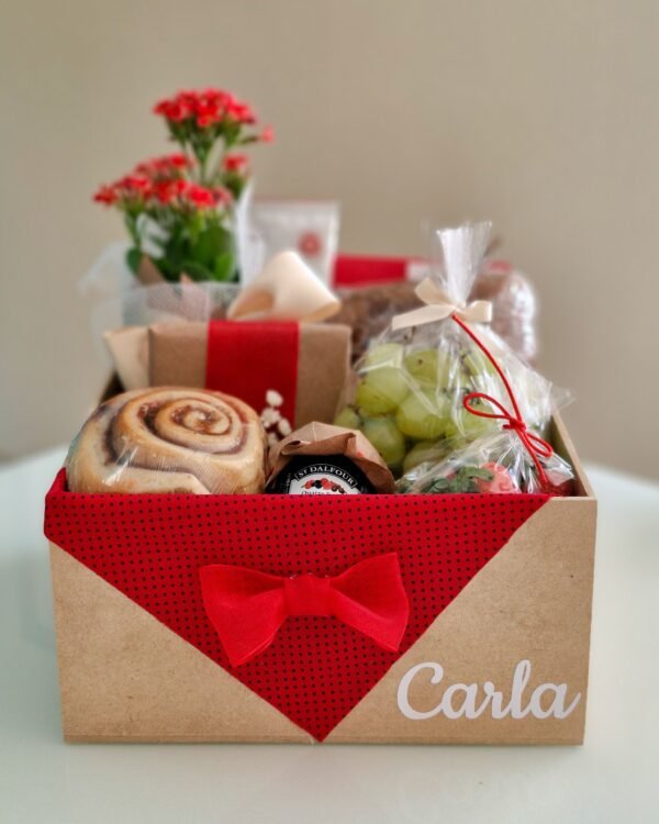 Cesta Fitness 1 Cesta de café da manhã saudável em caixa de MDF decorada com tecido vermelho de poá (bolinhas pretas), um delicado laço vermelho e o nome 'Carla'. A cesta contém pão 100% integral, rolinho de canela, geleia St. Dalfour, uvas verdes, morangos frescos, flor Kalanchoe vermelha e um presente embrulhado em papel kraft com laço vermelho.
