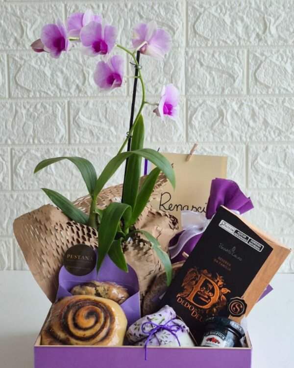 Cesta Orquídea 2 Detalhe frontal da cesta de café da manhã Orquídea, mostrando uma barra de chocolate premium escuro, rolinho de canela com calda brilhante, cookie artesanal no envelope roxo da Pestana, potinho decorado com amarração rústica e geleia. Ao fundo, as flores lilases da orquídea completam a composição sofisticada.