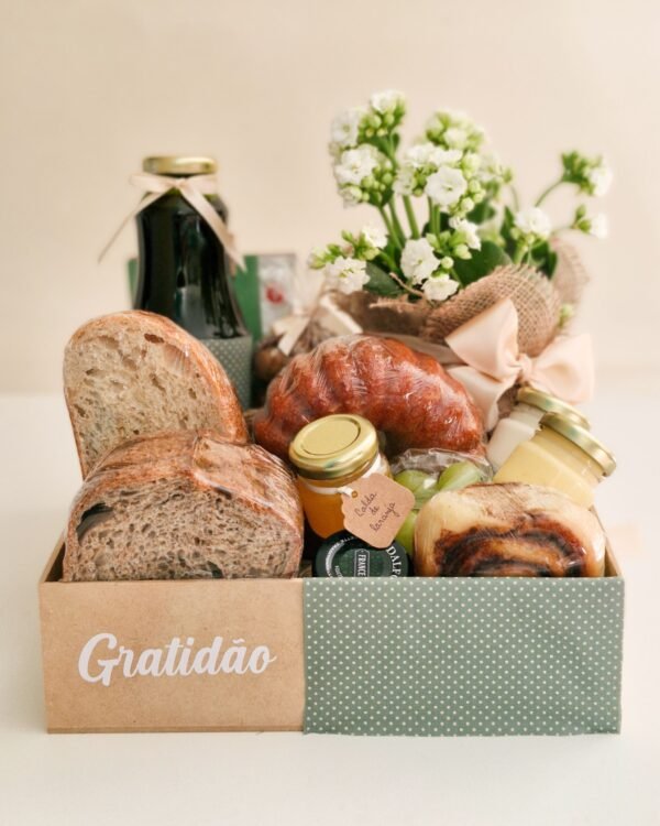 Cesta Gratidão 1 Cesta de café da manhã farta em caixa de MDF dividida entre acabamento cru com a palavra 'Gratidão' e tecido verde com bolinhas brancas (poá). O interior revela pães de fermentação natural, suculento bolo caseiro, calda de laranja artesanal em potinho de vidro, uvas verdes, suco, rolinho de canela e flor Kalanchoe branca.