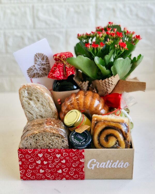 Cesta Gratidão 2 Cesta farta de café da manhã em caixa de MDF forrada com tecido vermelho estampado de corações brancos e a palavra 'Gratidão'. A composição inclui pães rústicos de fermentação natural, mini bolo caseiro, rolinho de canela, geleia St. Dalfour, flor Kalanchoe vermelha, suco em garrafa de vidro e um lindo cartão com coração de barbante.