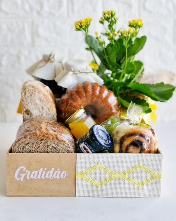 Cesta Gratidão 4 Cesta de café da manhã em caixa de MDF com a palavra 'Gratidão' e tecido bege texturizado com detalhes de costura em ziguezague amarelo. A composição exibe fatias grossas de pães rústicos, mini bolo caseiro, geleias, rolinho de canela, sucos em garrafas de vidro com tampas de tecido, uvas verdes e flor Kalanchoe amarela.