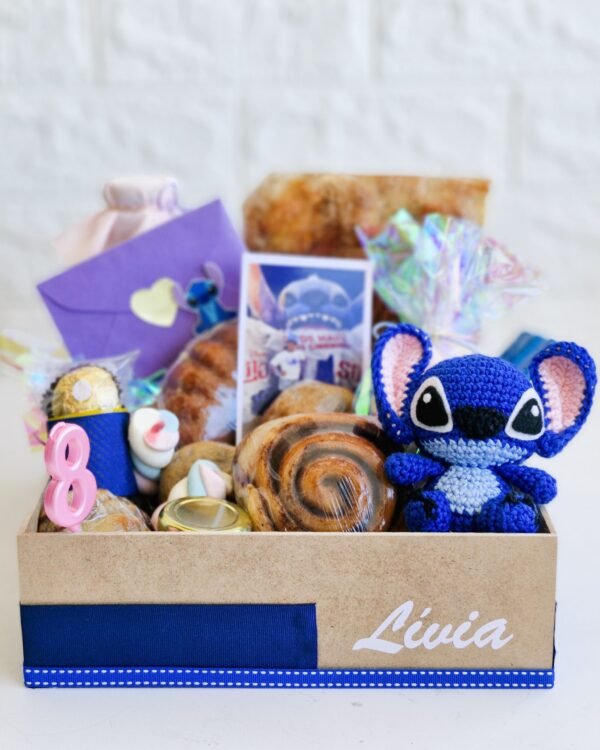 Inicio 4 Cesta de café da manhã infantil em caixa de MDF decorada com fita azul e o nome 'Lívia' em branco. Em destaque, um boneco amigurumi em crochê do personagem Stitch, uma foto revelada da criança, um envelope lilás temático, vela rosa de aniversário número 8, bombom Ferrero Rocher, marshmallows coloridos, rolinho de canela e pães artesanais rústicos
