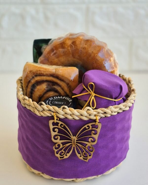 Cesta Momento Doce 2 Cesta de café da manhã em cesto de taboa envolto em tecido roxo vibrante, decorado com um aplique de borboleta de madeira vazada. A cesta farta exibe um mini bolo de laranja, rolinho de canela com calda, geleia St. Dalfour e um potinho de vidro com tampa de tecido roxo perfeitamente harmonizado.