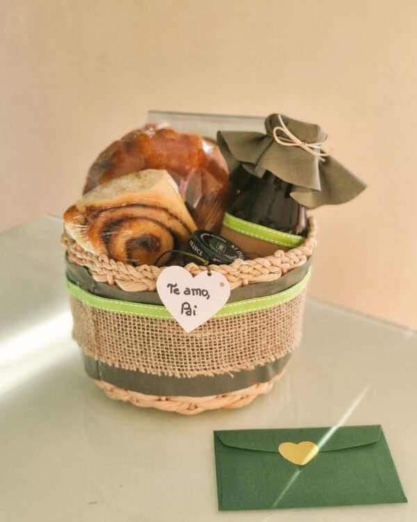 Cesta Momento Doce 1 Cesta rústica de taboa decorada com fita verde e faixa larga de juta, exibindo uma tag frontal em formato de coração com a frase 'Te amo, Pai' escrita à mão. No interior: rolinho de canela, mini bolo caseiro, geleia St. Dalfour e uma garrafinha com tampa de tecido verde musgo. Em primeiro plano, um envelope verde com selo de coração dourado.