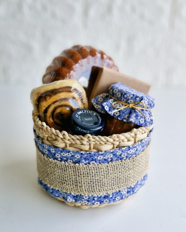 Inicio 3 Cesta de café da manhã em cesto de taboa decorado com tecido floral azul e faixa de juta rústica. A composição farta contém um mini bolo de laranja caseiro, rolinho de canela macio, geleia francesa St. Dalfour, caixa kraft e um potinho de vidro com tampa de tecido floral azul combinando com a embalagem.