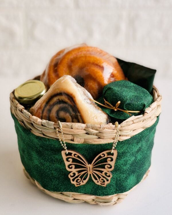 Cesta Momento Doce 4 Cesta de café da manhã em cesto rústico de taboa, inteiramente revestido com tecido verde esmeralda texturizado e decorado com um elegante aplique de borboleta de madeira pendurado por uma correntinha dourada. O interior farto contém mini bolo de laranja, rolinho de canela, geleia e um recipiente com tampa de tecido verde combinando.