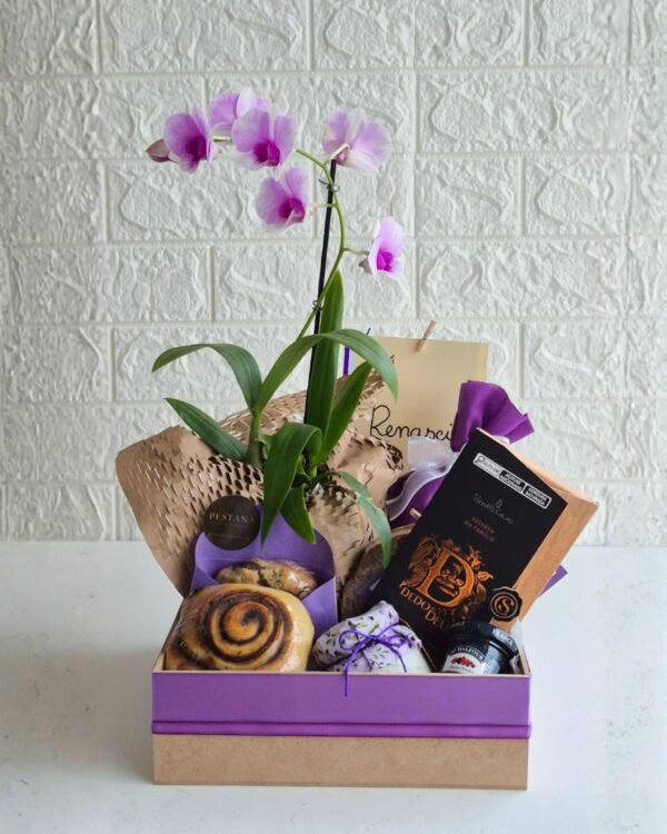 Inicio 2 Cesta de café da manhã premium em caixa de MDF com base crua e tampa roxa. Em destaque, uma orquídea lilás alta envolvida em papel kraft sustentável favo de mel. A composição inclui barra de chocolate Dedo de Deus, rolinho de canela, cookie artesanal em envelope roxo com selo Pestana, potinho de pasta salgada com tampa de tecido floral roxo, geleia francesa St. Dalfour e cartão.