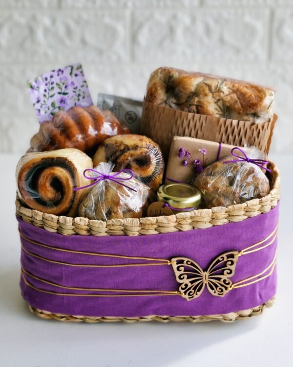 Cesta Taboa 3 Cesta de café da manhã em cesto rústico de taboa decorado com tecido roxo, fios dourados e aplique de borboleta em madeira. No interior farto, focaccia de fermentação natural em embalagem kraft, mini bolo caseiro, rolinhos de canela, cookies embalados com laço roxo, potinho de vidro e cartão floral.