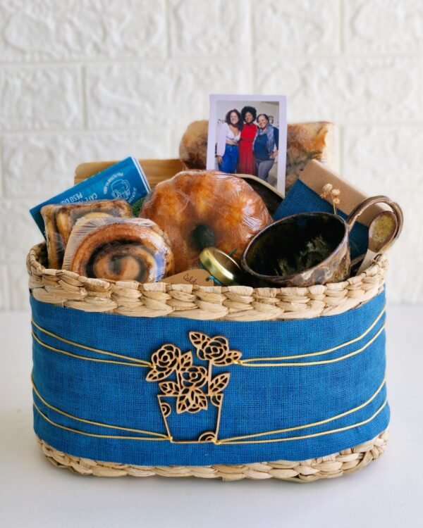 Cesta Taboa 2 Cesta de café da manhã rústica em cesto de taboa decorado com tecido azul, fios dourados e aplique de vaso de flores em madeira. A composição destaca uma elegante xícara de cerâmica artesanal com colher de madeira, uma foto revelada de família, mini bolo de laranja, rolinhos de canela, drip coffee, potinho de vidro e focaccia de fermentação natural.