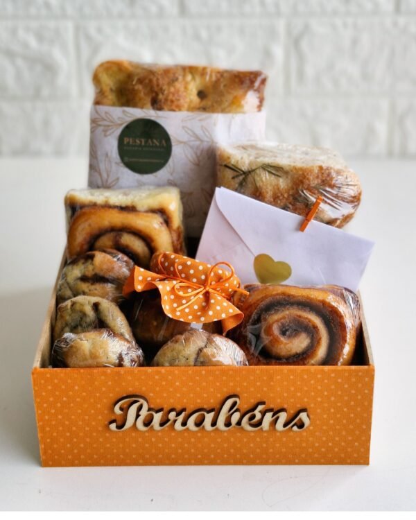 Inicio 5 Cesta de café da manhã de aniversário em caixa laranja com a palavra "Parabéns" em madeira. Contém pães artesanais, biscoitos, geleia e um cartão com coração.