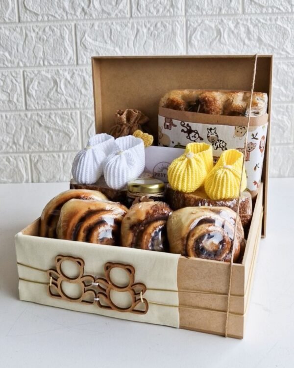 Cesta Boas Vindas 3 Cesta de Boas-Vindas maternidade grande em caixa kraft decorada com faixa de tecido creme e aplique de dois ursinhos de madeira vazados. A cesta é farta, contendo quatro rolinhos de canela, pão rústico com embalagem de safari, geleia, um saquinho de juta, cartão da Pestana e o grande destaque: dois pares de sapatinhos de crochê, sendo um branco e um amarelo, arranjados sobre os pães.