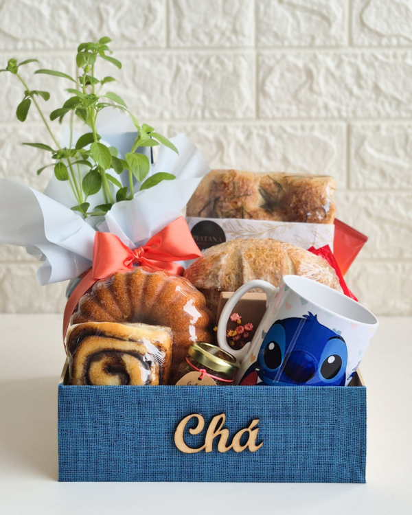 Cesta Chá da Tarde 1 Cesta de chá da tarde ou café da manhã em caixa azul texturizada com a palavra "Chá" em madeira. A cesta contém uma caneca personalizada do personagem Stitch (Disney), um vaso de manjericão fresco embalado para presente com laço laranja, pães rústicos de fermentação natural, um bolo caseiro redondo, rolinho doce e um pote de vidro pequeno (geleia/mel).