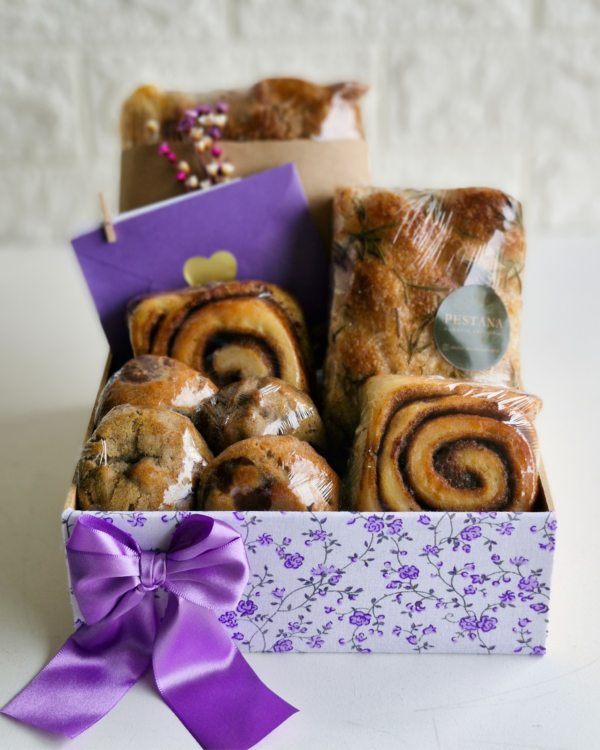 Cesta Encanto 8 Cesta de café da manhã Encanto em uma delicada caixa com estampa floral lilás e um vistoso laço de cetim roxo. Acomoda pães rústicos de fermentação natural decorados com alecrim (um com adesivo da Pestana), suculentos rolinhos de canela (cinnamon rolls), cookies com gotas de chocolate, e um envelope roxo selado com coração dourado.